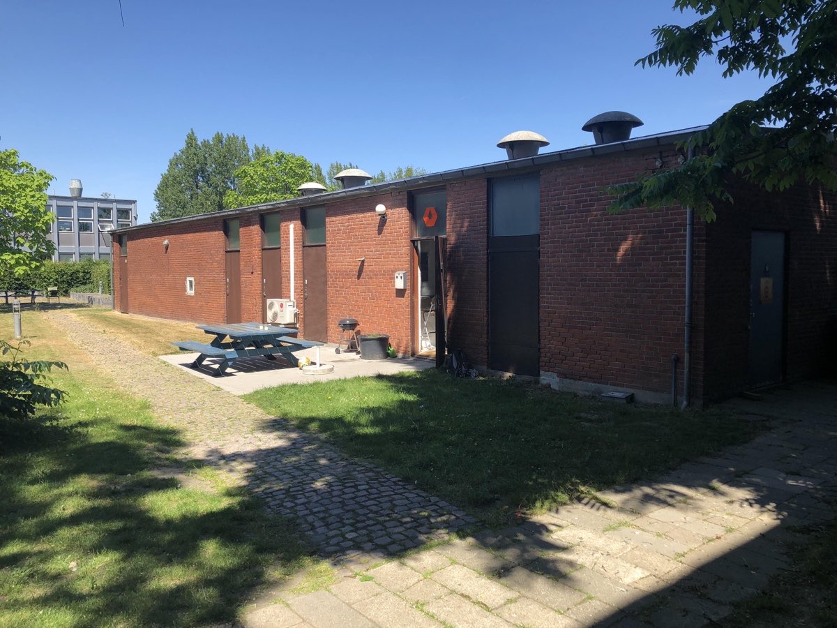 Facaden på Raffinaderivej i solskin — rød murstensbygning med gårdareal og bordbænkesæt